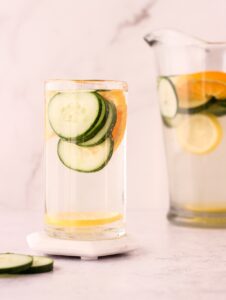 Glass of water with fruit and veggie slices in it in front of a pitcher of water