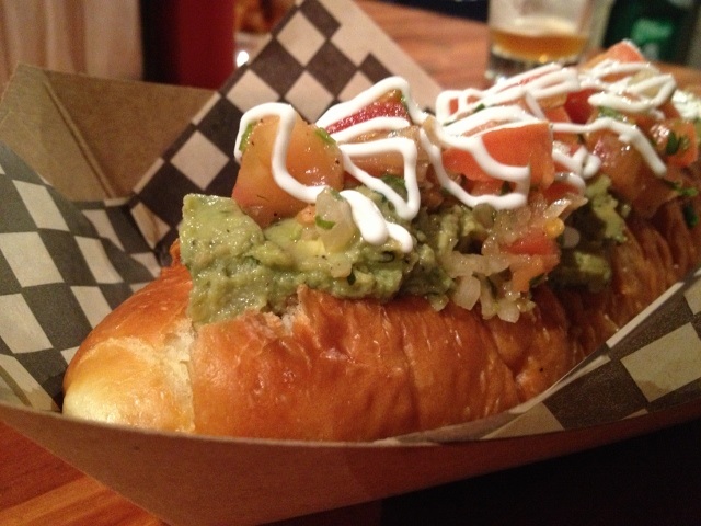 Hot dog in a paper tray with guacamole and tomatoes