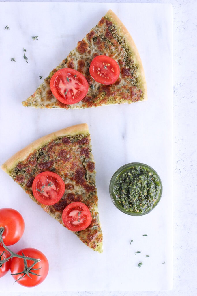 Top down view of 2 slices of pizza on a marble cutting board with a jar of pesto