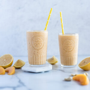 Two glasses of peach smoothie surrounded by peaches and lemon slices