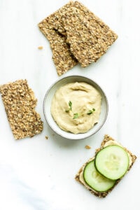Bowl of hummus next to crackers topped with cucumber slices