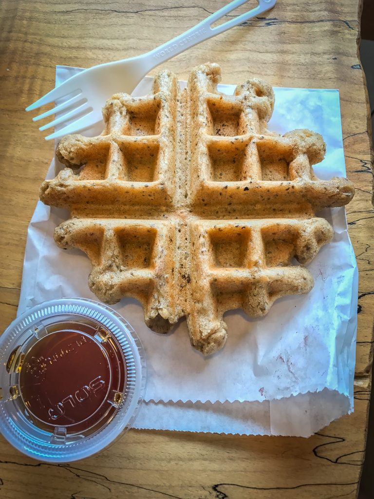 A small waffle sitting on a paper sack, alongside a fork and a container of maple syrup