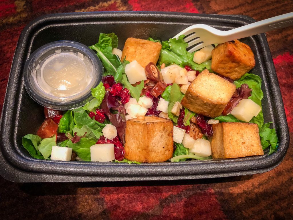 A black plastic to-go container filled with greens, tofu, dried fruit and a container of dressing