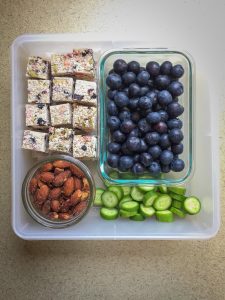 Square plastic container with blueberries, coconut bites, cucumber slices and almonds