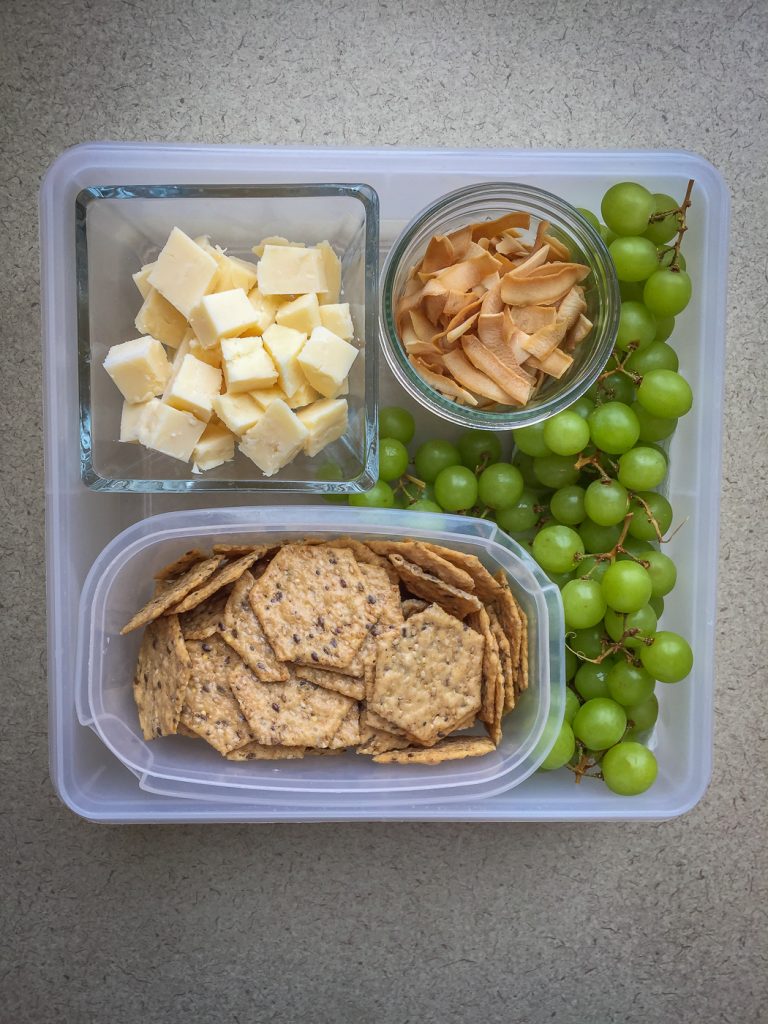 Plastic container with cheese, coconut chips, grapes, and multigrain crackers