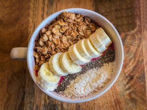 White ceramic bowl filled with red smoothie and topped with granola, coconut, banana, and chia seeds