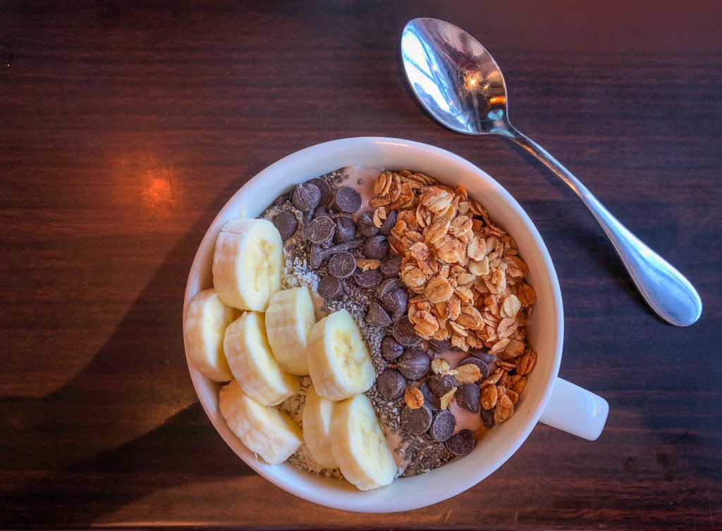 Large white mug filled with smoothie topped with banana, chia seeds, chocolate chips and granola next to a spoon