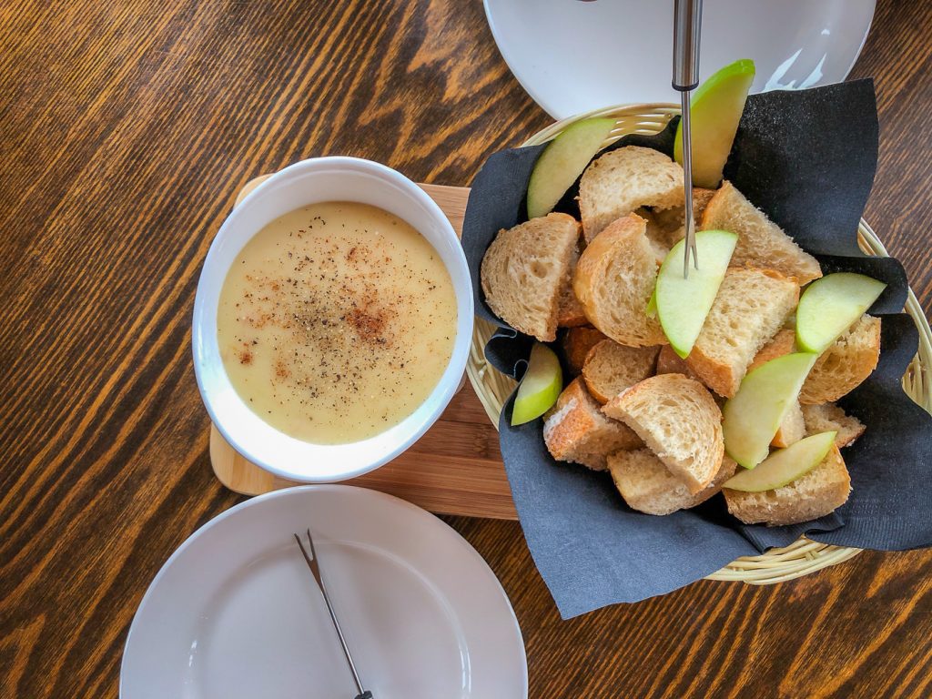 Basket of bread and apple slices beside a small white pot of cheese fondue