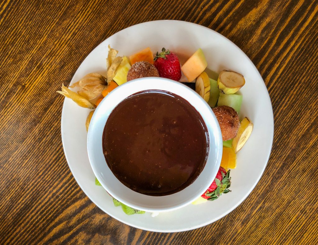 Top down photo of a plate of fruit and donut holes surrounding a fondue pot of chocolate