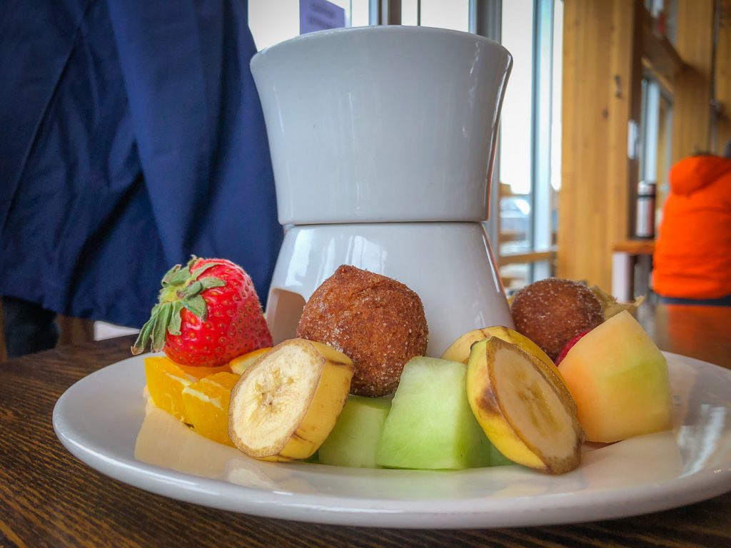 Side shot of a small pot of chocolate fondue surrounded by banana slices, berries, and melon