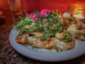 Thick slice of roasted cauliflower on a plate topped with green salsa and pickled onions