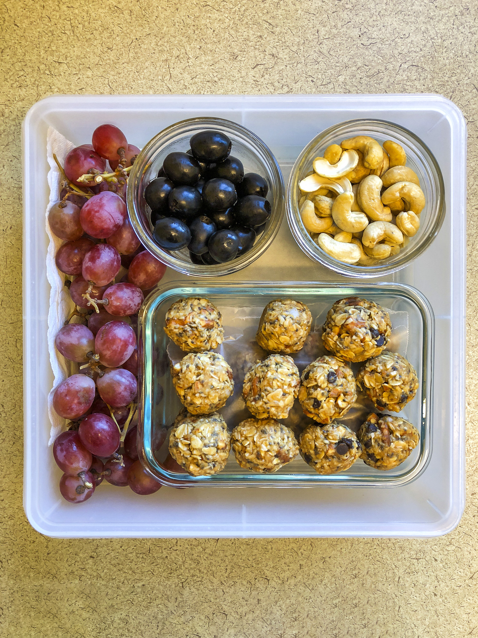 Tupperware filled with olives, cashews, energy bites and grapes
