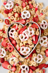 Close up on a heart shaped pan filled with red and white snack mix surrounded by more snack mix