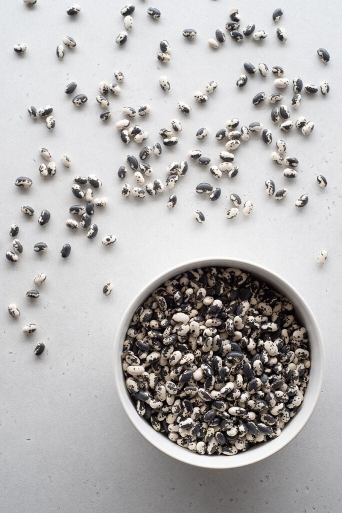 White bowl filled with black and white speckled beans next to some beans scattered on a white surface