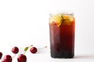 Large glass of dark cherry vinegar in fizzy water garnished with lime surrounded by a handful of scattered cherries