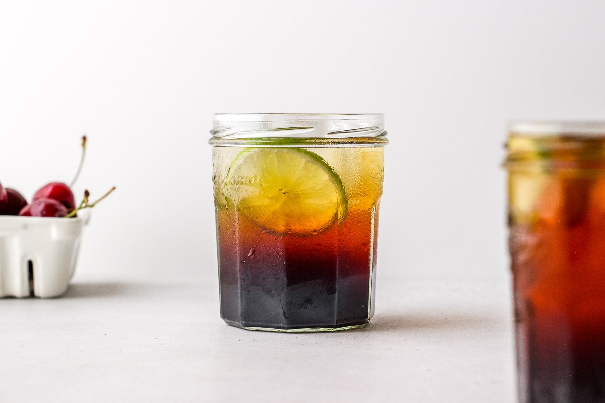 Small glass filled with dark vinegar and fizzy water with lime garnish on white surface next to a second glass and a basket of cherries