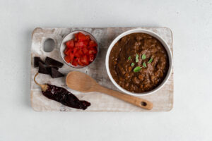 Tope down view of a cutting board topped with chiles, tomatoes, chocolate and a bowl of mole
