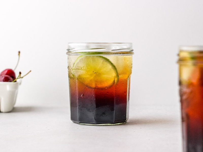 Small glass filled with dark vinegar and fizzy water with lime garnish on white surface next to a second glass and a basket of cherries